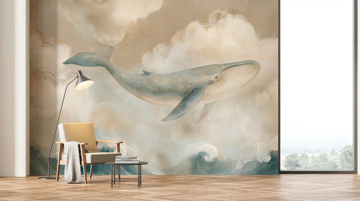 LINEN WHALE en toda la pared de un salón con ventanal: ballena y nubes cremosas sobre un sillón con lámpara de pie, olas tras una mesita baja